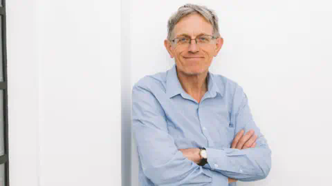 一位戴眼镜、穿着牛仔裤和蓝色敞开领衬衫的灰发男子，笑容满面，靠在白色墙壁上，右侧是一扇老式工业风格的窗户。