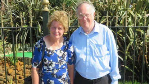 家庭资料：Lois and John McCullough，他们站在一个迷你高尔夫球场前。他们穿着蓝色衣服，对着摄像头微笑。